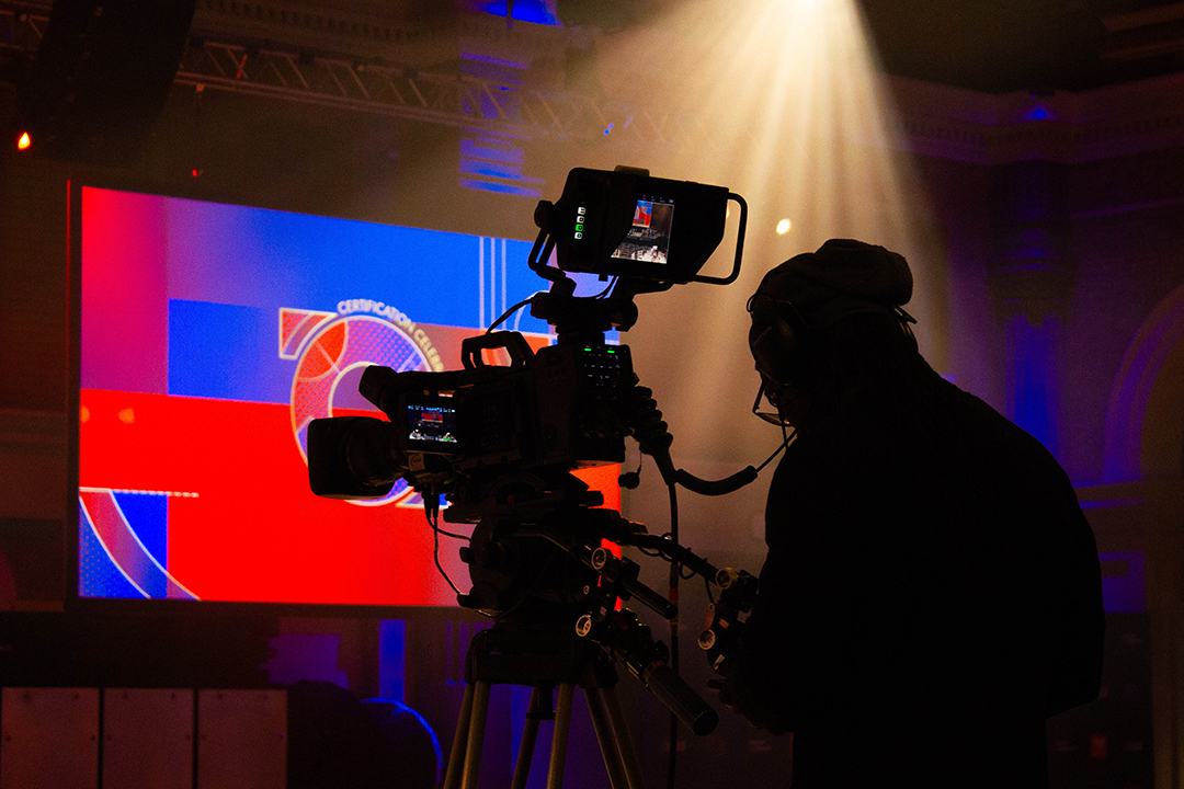 A silhouette of a camera operator filming indoors, with bright lights overhead and a colorful screen displaying Top Employer Awards graphics in the background.