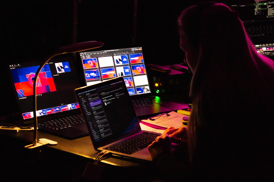 A person working at a dark desk with multiple laptop and monitor screens displaying colorful graphics, illuminated by soft light, possibly managing a Top Employer Awards live event or broadcast.