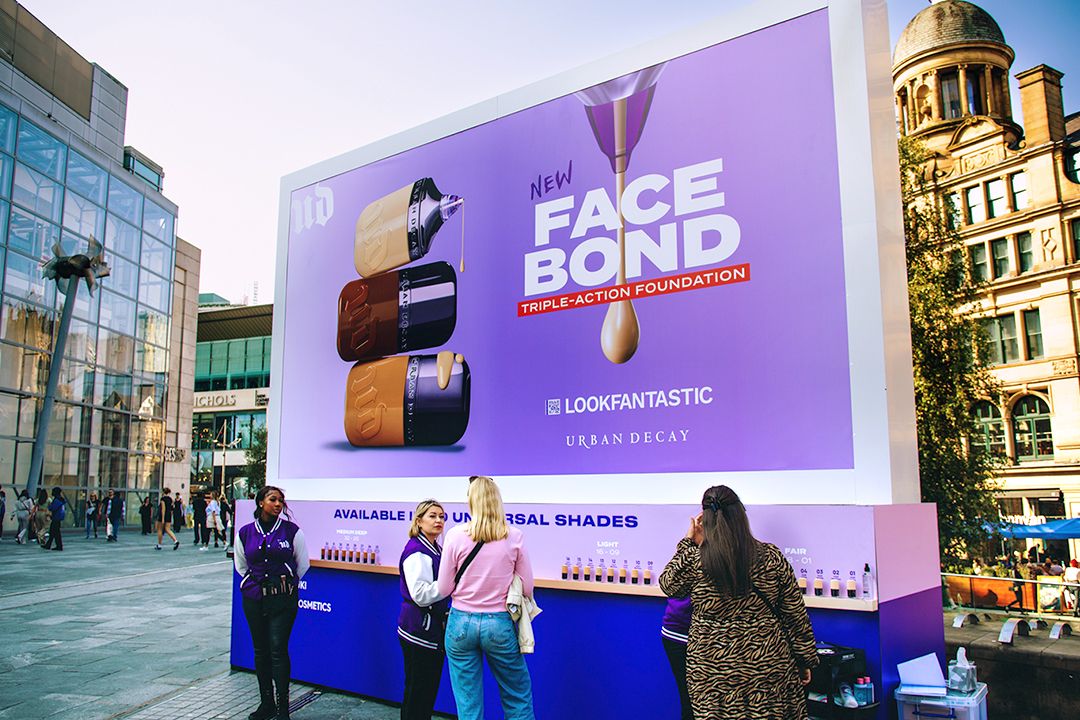 A large outdoor billboard advertises the new Urban Decay Face Bond foundation, showcasing three bottles and product swatches. Several people gather around a Face Bond display below the ad in a busy city square.