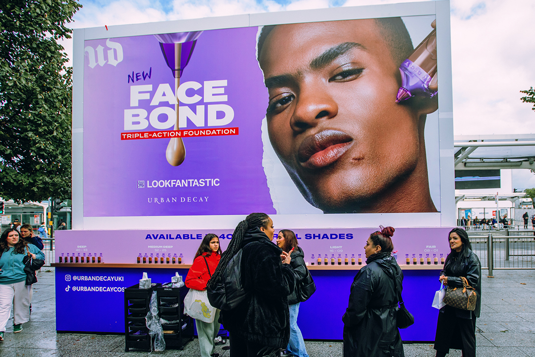 A large outdoor billboard advertises Urban Decay Face Bond foundation, featuring a close-up of a model’s face. Several people stand and interact in front of the display, highlighting the new Urban Decay Face Bond bottles.