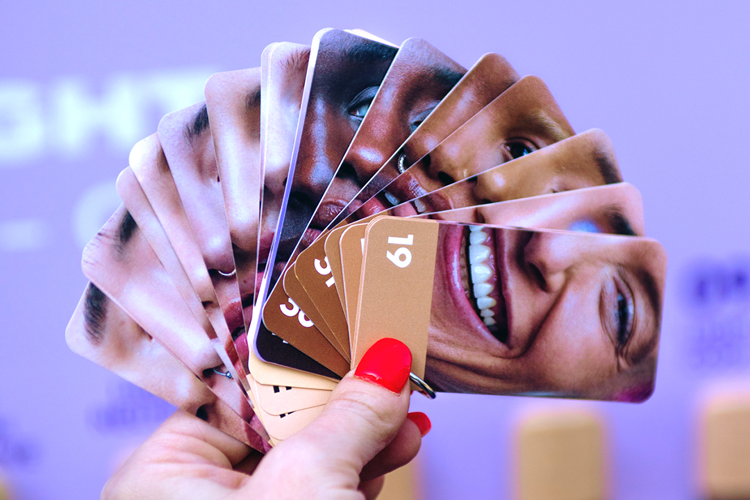 A hand with red nail polish holds a fan of cards, each displaying a different skin tone and facial feature, against a blurred purple background—a nod to Urban Decay Face Bond’s celebration of diversity.