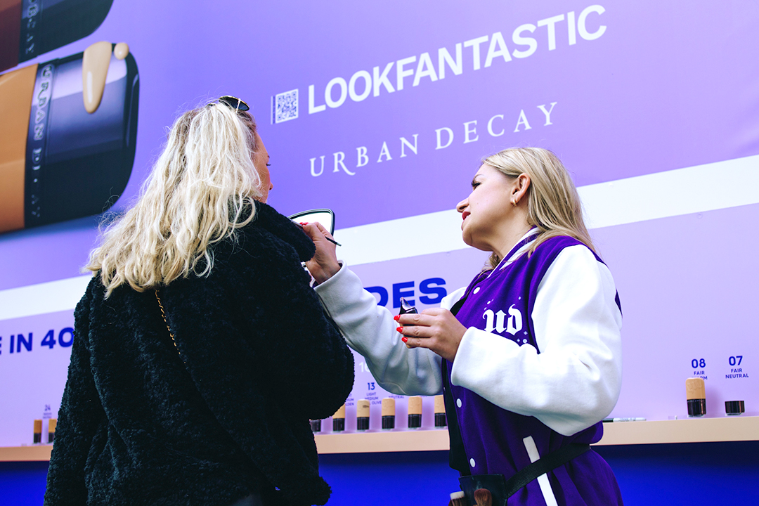 A makeup artist applies makeup to a woman in front of a purple Urban Decay and LOOKFANTASTIC display featuring various foundation shades, highlighting the new Urban Decay Face Bond for a flawless finish.