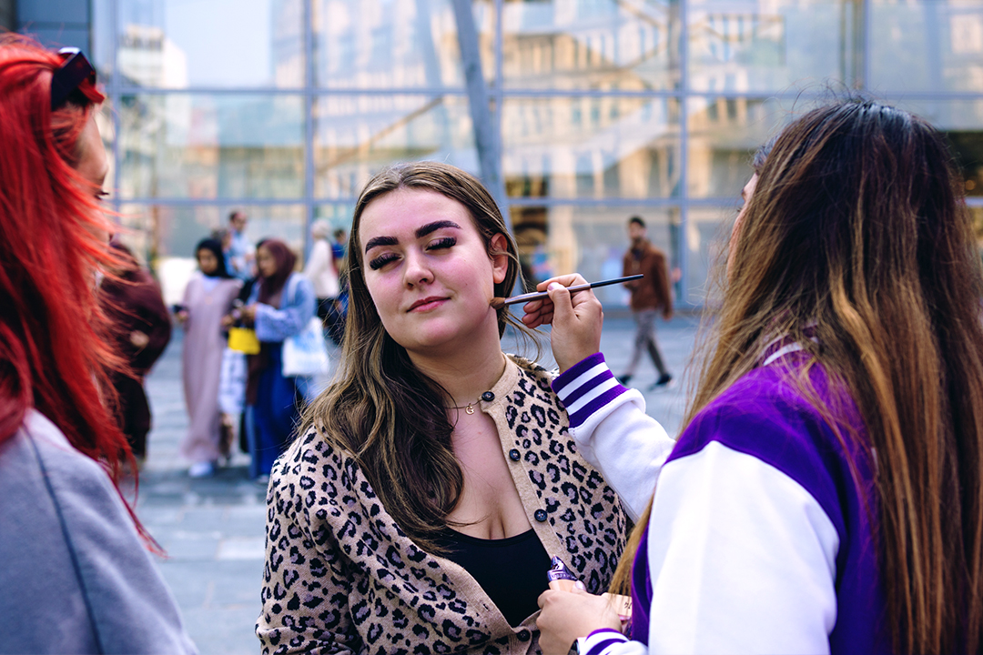 A woman with long brown hair and a leopard-print jacket is having her makeup done with Urban Decay Face Bond by another woman in a purple and white jacket, outdoors in an urban setting with people walking in the background.