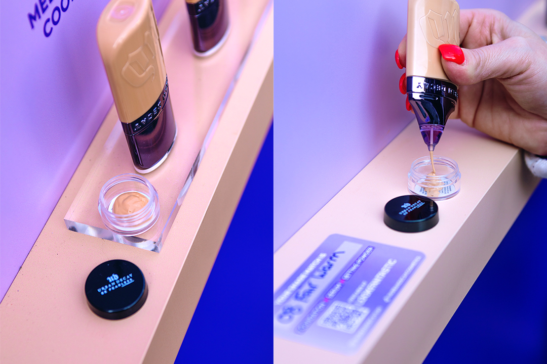A close-up of a hand dispensing liquid foundation, like Urban Decay Face Bond, from a bottle into a small clear container, with makeup bottles and a black lid nearby on a beige and purple surface.
