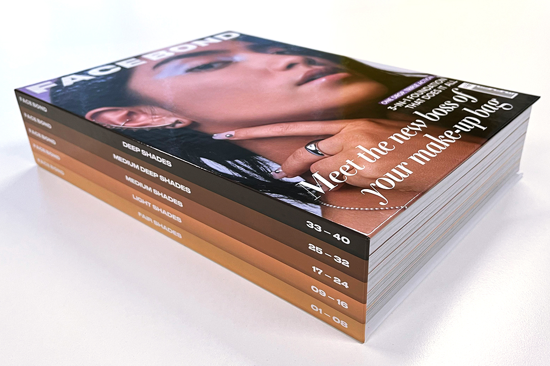 A stack of Urban Decay Face Bond magazines with a cover showing a close-up of a person’s face and hand. Text on the cover says, “Meet the new boss of your make-up bag.” The spines are labeled by shade ranges.