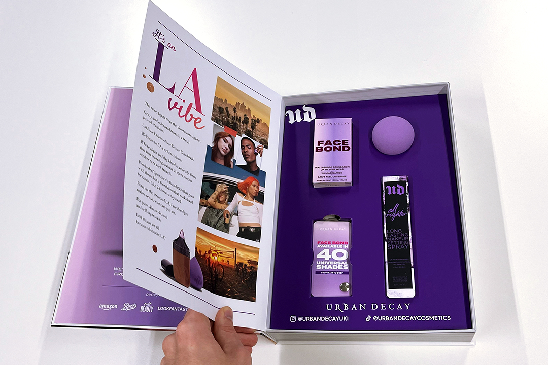A person holds open a box containing Urban Decay cosmetics, featuring Urban Decay Face Bond foundation, All Nighter setting spray, and a purple makeup sponge. The inside cover displays colorful photos and product information.