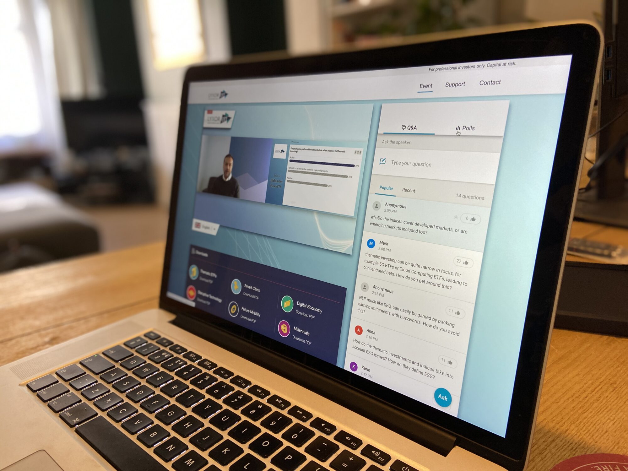 A laptop on a wooden desk displays a virtual event featuring the Lyxor ETF case study, with a speakers video and a chat panel showing questions and comments. The background is a cozy, blurred indoor setting.