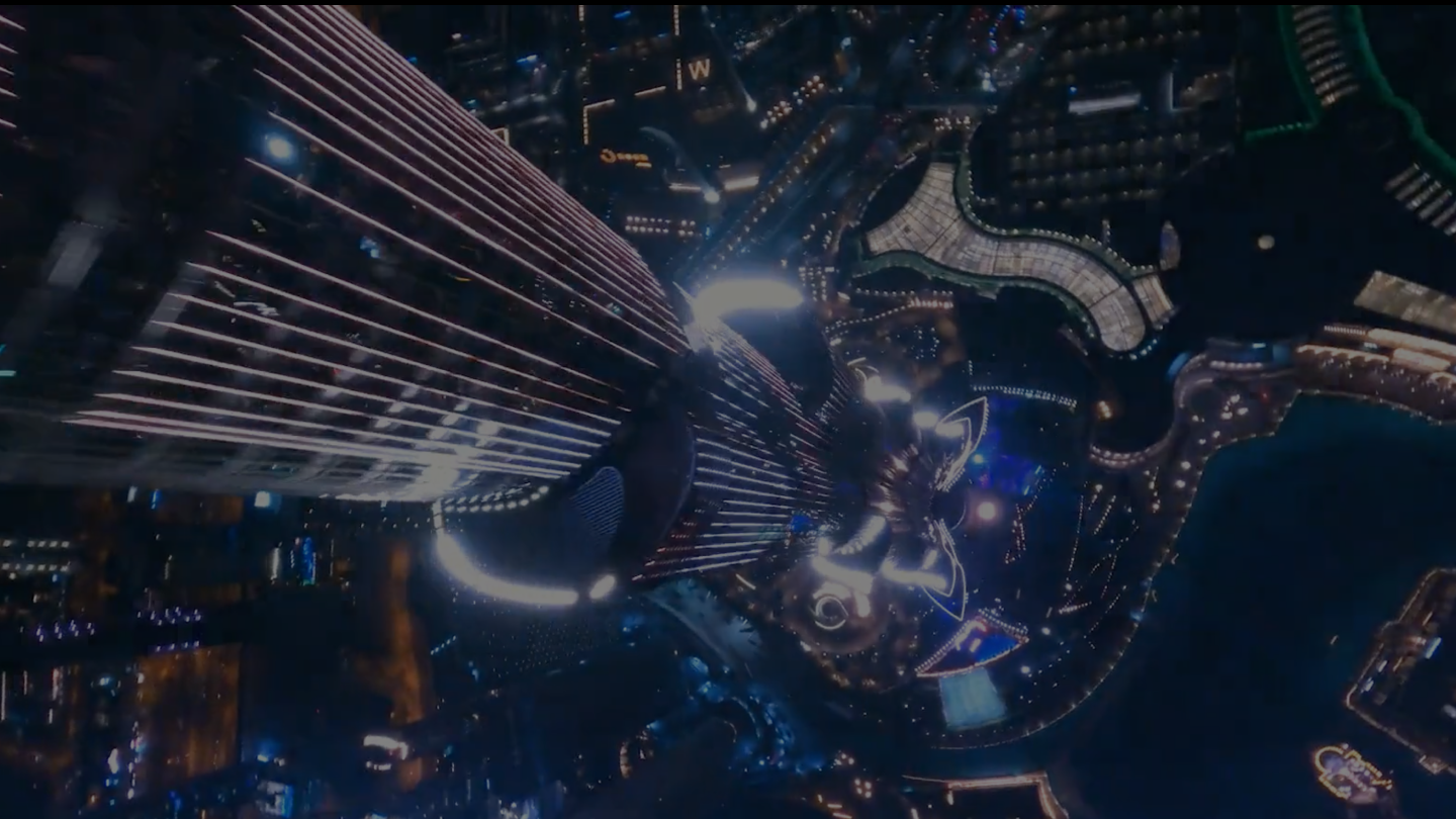 A nighttime aerial view looking down the side of a brightly lit skyscraper, surrounded by illuminated city streets and fountains—a scene reminiscent of an immersive marketing agency’s creative vision.