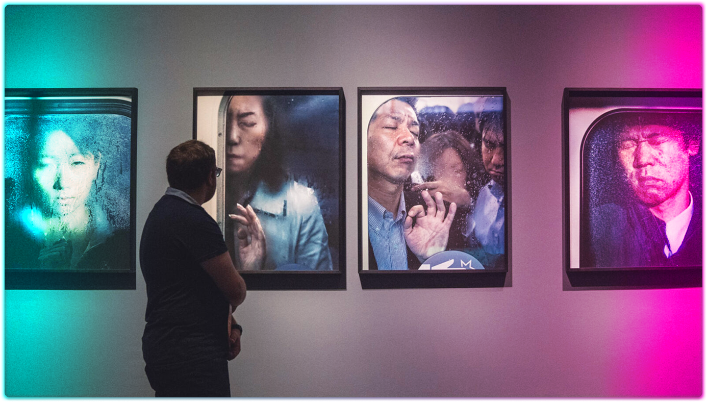 A person stands in front of four framed photographs on a gallery wall, each photo showing people pressed against glass, appearing crowded and contemplative—an immersive marketing approach highlighted by colorful pink and blue lights.