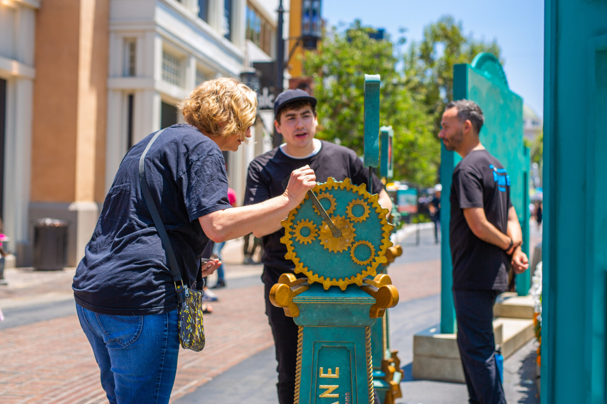 A woman turns a large decorative gear on an Arcane FYC Emmys street installation, while two men in black shirts observe. The scene is outdoors on a sunny day, with buildings and trees in the background.