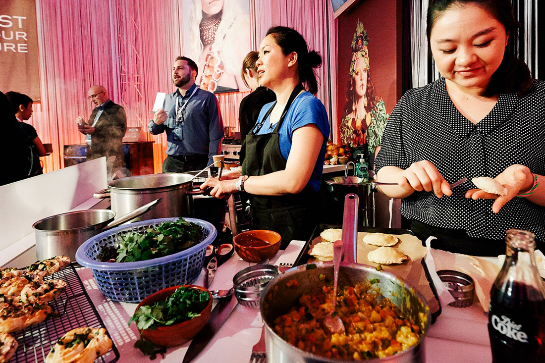 Three people prepare and cook food at a busy indoor food stall. Various ingredients and dishes are on the counter, resembling a Beko case study, as chefs focus intently amid the lively crowd mingling in the background.