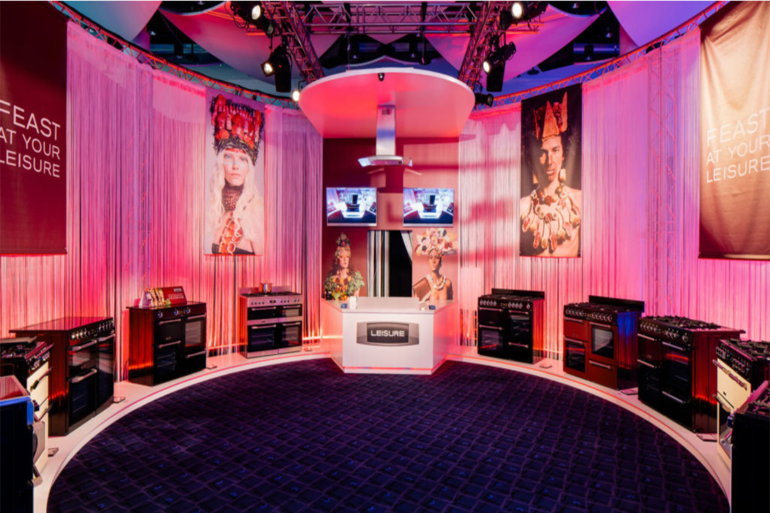 A circular showroom with stoves and ovens displayed along curved walls, dramatic pink lighting, and large banners of people in ornate headdresses. Inspired by the Beko case study, a central counter with LEISURE branding sits under a modern canopy.