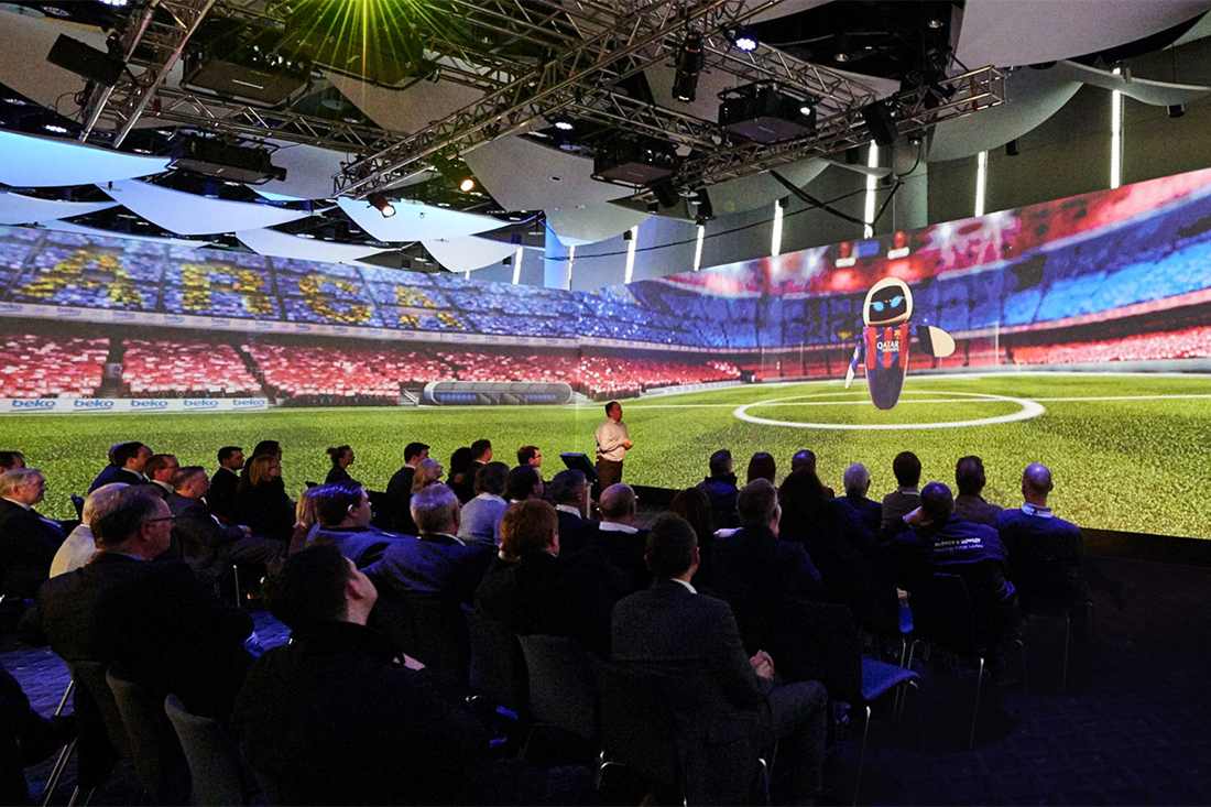 A group of people sit in a darkened auditorium watching a large screen displaying a soccer stadium with a cartoon robot mascot, as stage lighting and technical equipment glow overhead during the Beko case study presentation.