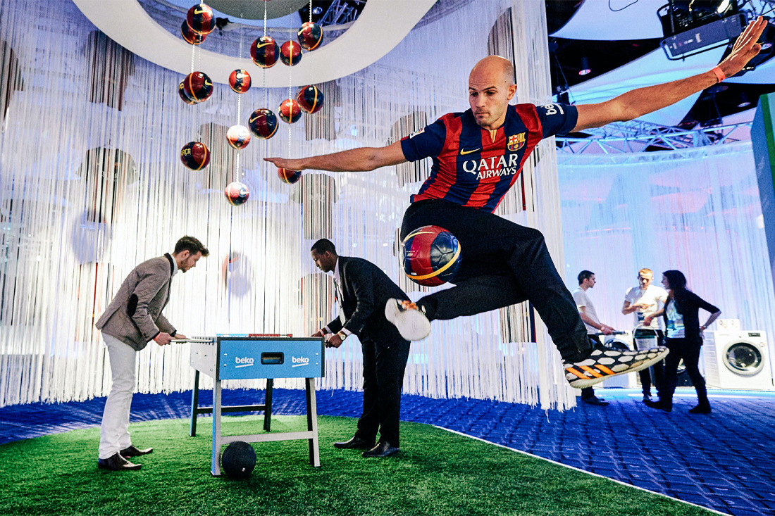 A man in a Barcelona soccer jersey jumps mid-air, kicking a ball, while two others play foosball nearby—capturing the lively spirit often highlighted in a Beko case study. Hanging soccer balls and white streamers decorate the modern indoor setting.