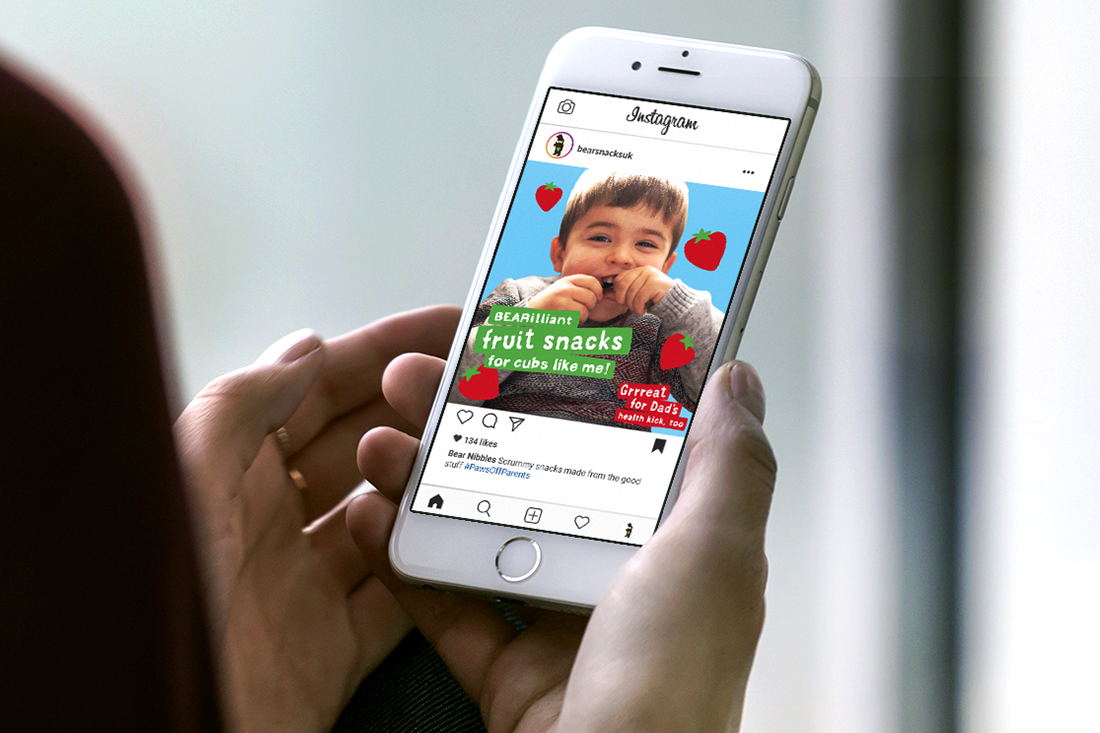 A person holding a smartphone displaying an Instagram post featuring a smiling child with hands under their chin, surrounded by red hearts. The post advertises BEAR fruit snacks with the slogan, “fruit snacks for cubs like me!”—a Bear Nibbles case study.