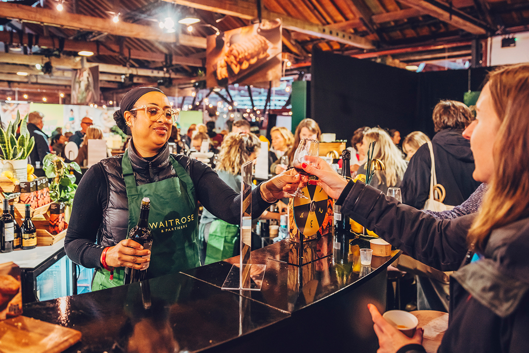 A woman wearing a green Waitrose apron smiles as she serves a drink to an attendee at the bustling Waitrose Winter Festival, with people and stalls visible in the background.