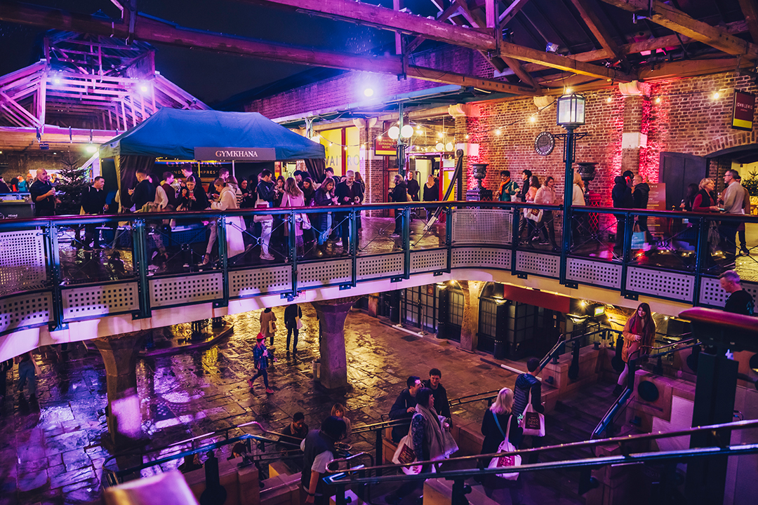 A vibrant indoor market scene at night during the Waitrose Winter Festival, with people walking on an upper-level walkway and gathering below, illuminated by colorful purple and yellow lights against exposed brick walls and wooden beams.