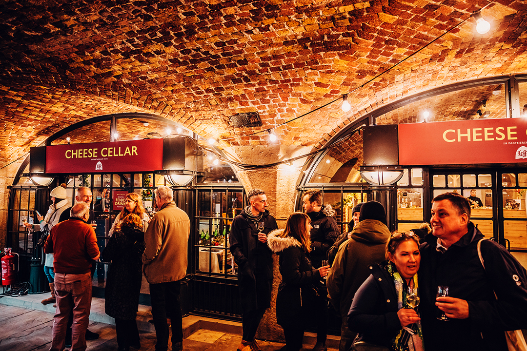People gather and socialize inside a warmly lit, brick-ceilinged cheese cellar at the Waitrose Winter Festival, holding drinks and chatting near arched displays stocked with bottles and cheeses.