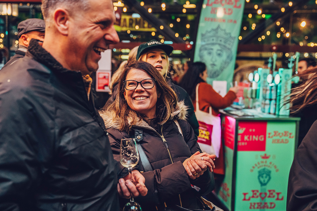 A group of people smiling and laughing at an indoor Waitrose Winter Festival event. A woman in glasses holds a wine glass, while a man beside her laughs. Festive lights and a colorful bar booth brighten the background.