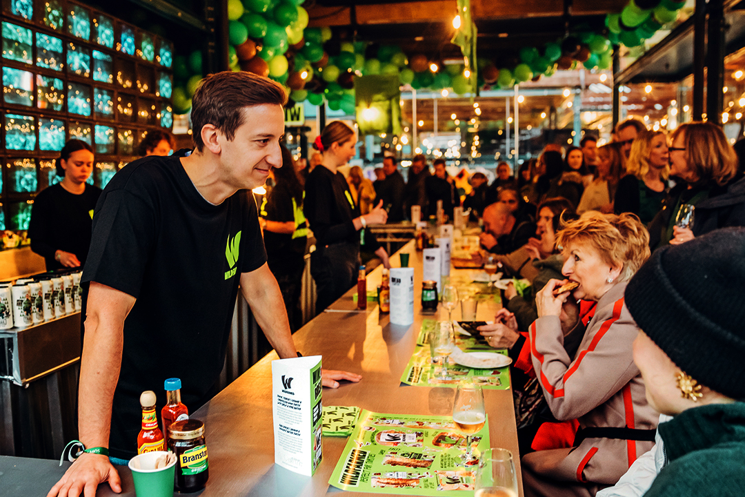 A male server in a black shirt talks to seated customers at a lively, crowded bar decorated with green balloons and lights, capturing the festive spirit of the Waitrose Winter Festival as people eat, drink, and socialize.