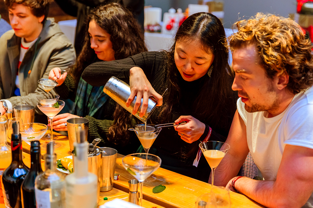 At the Waitrose Winter Festival, a group of people sit at a wooden table making cocktails, with one woman pouring a drink from a shaker into a martini glass while others watch and smile. Bottles and bar tools are scattered on the table.