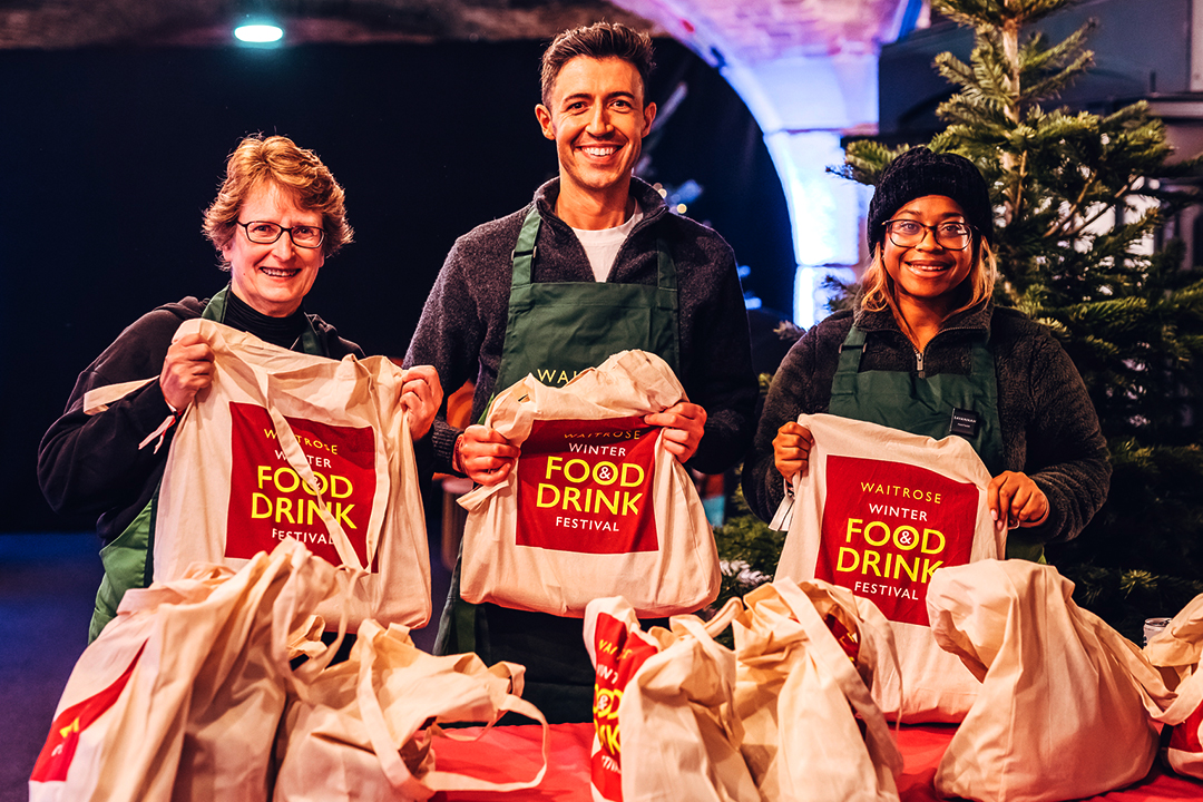 Three smiling people in green aprons stand behind a table, packing gift bags labeled Waitrose Winter Festival in a festive setting with a Christmas tree twinkling in the background.
