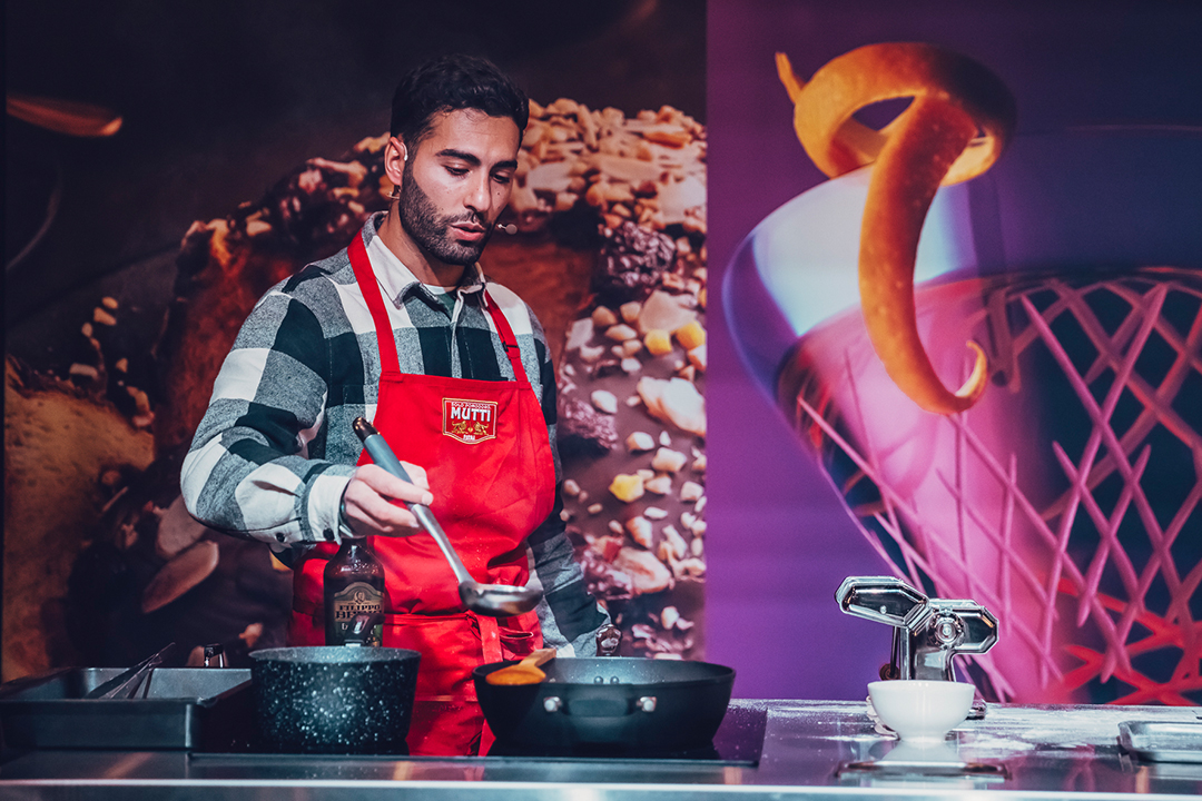 A man in a red apron cooks at a kitchen counter during the Waitrose Winter Festival, ladling food into a pan. Behind him is a large, colorful backdrop featuring food and a basketball net, with utensils and ingredients scattered on the counter.