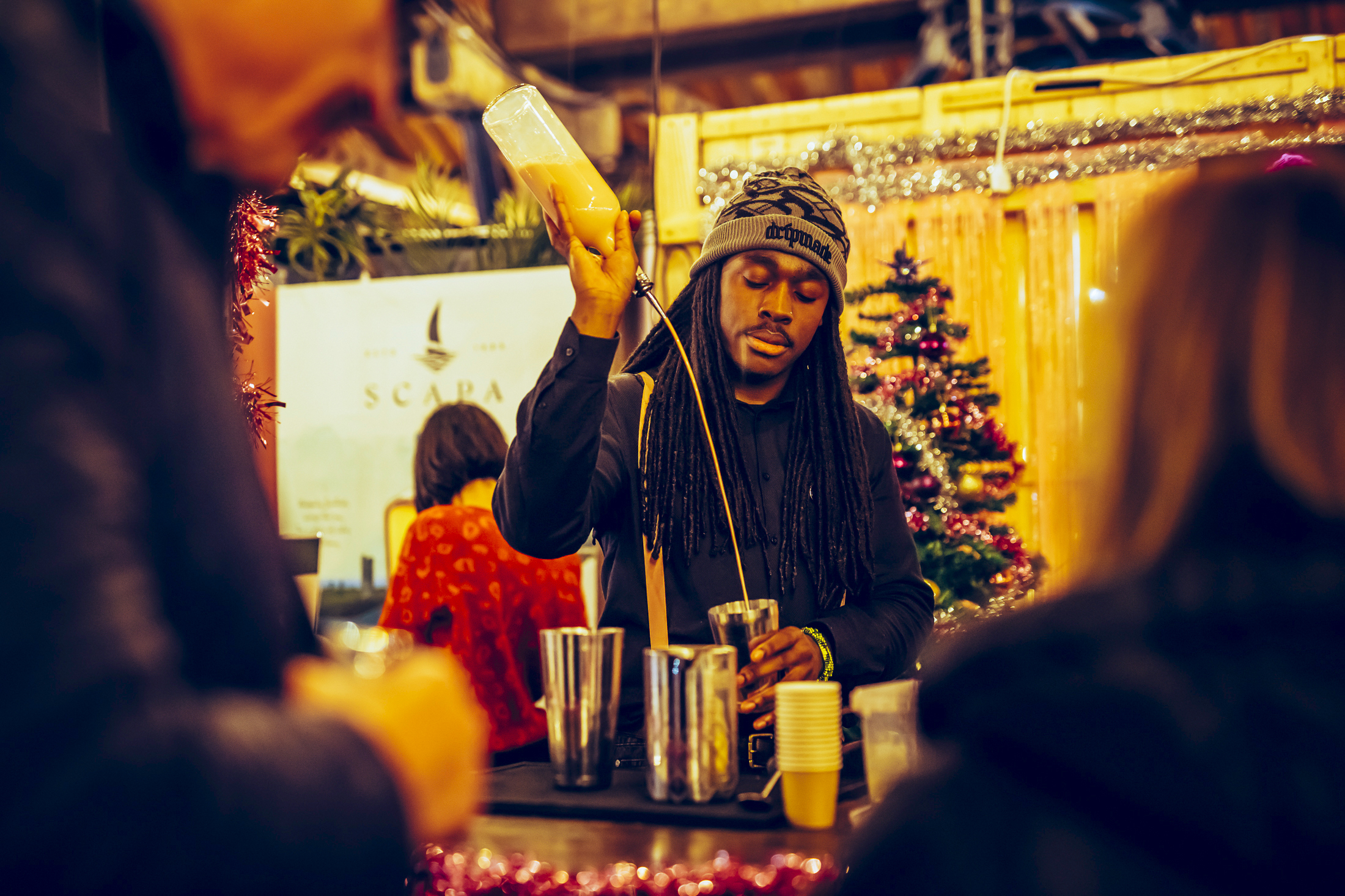 A bartender in a beanie pours a drink into a shaker at the Waitrose Winter Festival market stall, decorated with tinsel, with a Christmas tree in the background and people gathered around the counter.