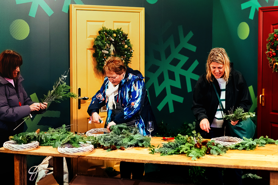 Three women craft holiday wreaths at a festive table adorned with greenery and pinecones. Two doors with Christmas wreaths and a large snowflake pattern set the scene for a creative Waitrose Winter Festival gathering.