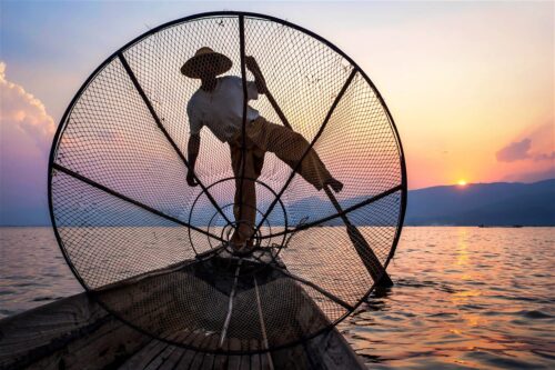 A fisherman stands on a wooden boat, balancing with one leg on a traditional fishing net frame—much like a marketing team working in harmony—silhouetted against a colorful sunset over calm water and distant mountains.