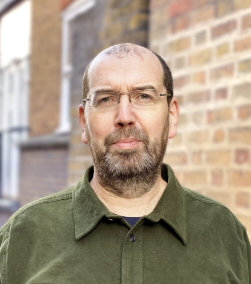 A middle-aged man with glasses, a beard, and short hair, wearing an olive green shirt, stands outdoors in front of a brick wall and blurred urban background—an ideal fit for any marketing team.