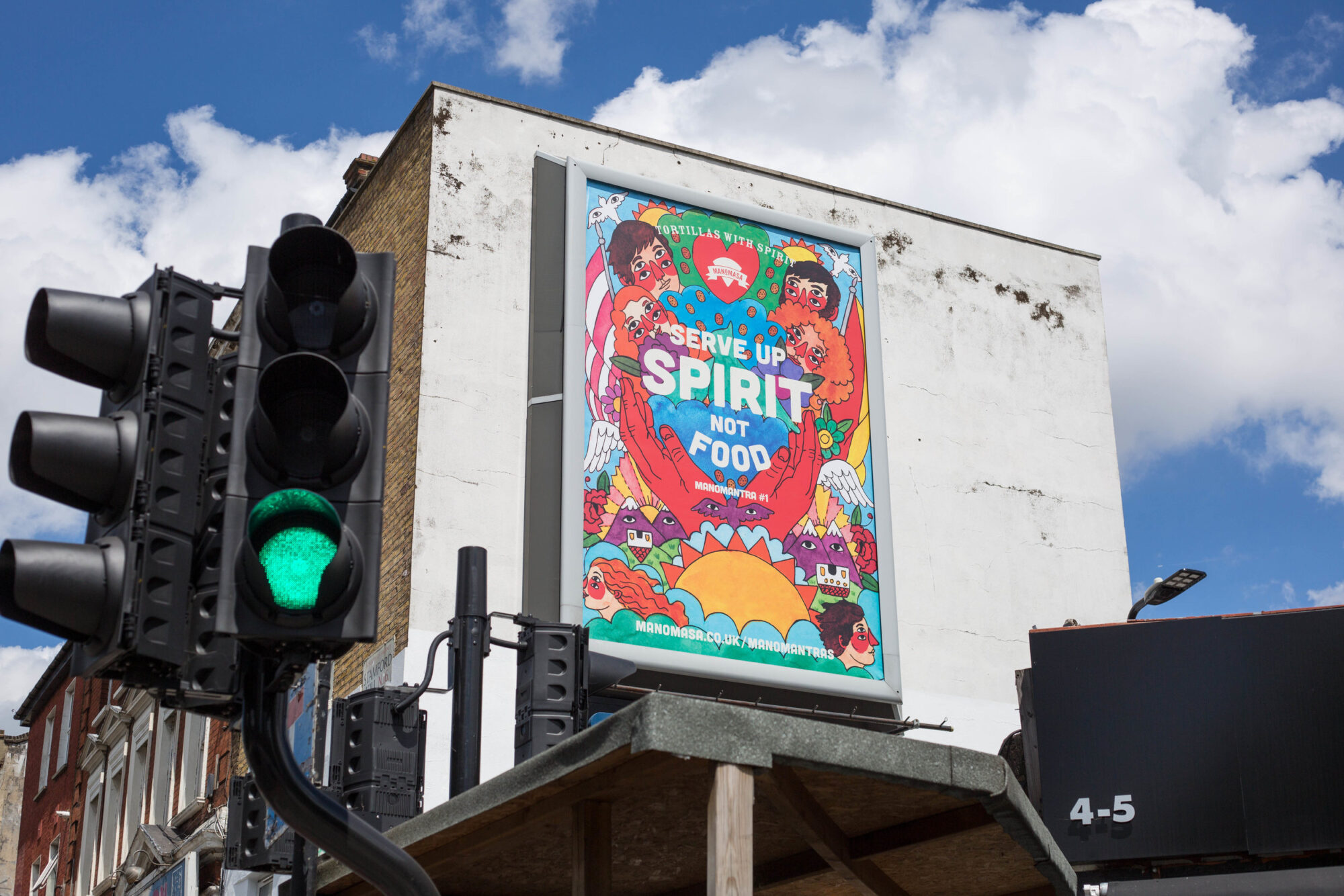 A colorful billboard on a building reads Serve Up Spirit Not Food, featuring illustrated people, hearts, and vibrant patterns—a scene reminiscent of a Manomasa case study. A green traffic light stands in the foreground under a partly cloudy sky.