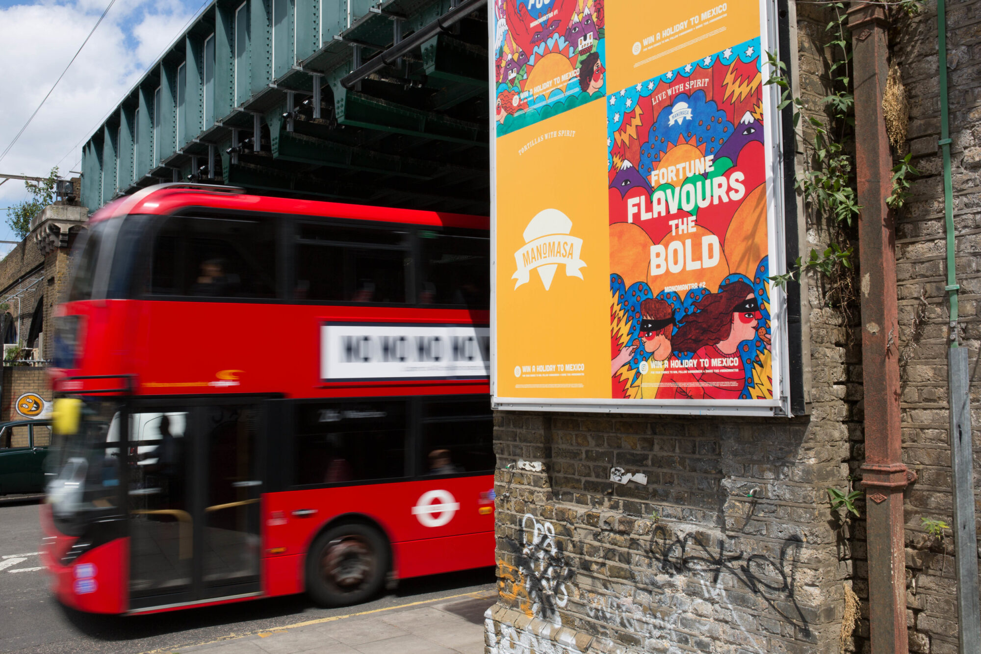 A red double-decker bus passes under a railway bridge next to a brick wall filled with colorful posters, including one from the Manomasa case study that says Fortune Flavours The Bold. The vibrant urban scene features graffiti on the wall.
