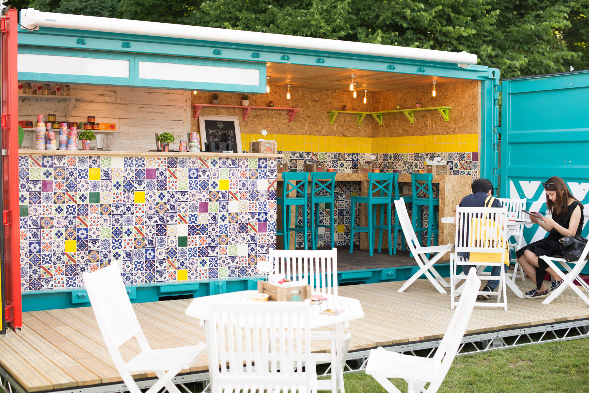 A colorful outdoor café made from a shipping container, decorated with patterned tiles, blue chairs, and stools; inspired by the Manomasa case study, two people sit at a white table on a grassy area, surrounded by more white tables and chairs.
