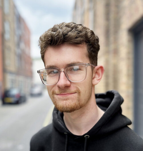 A young man with short brown hair, a trimmed beard, and clear glasses—dressed in a black hoodie—stands on a blurred urban street with brick buildings, looking like he just left a meeting with the marketing team.