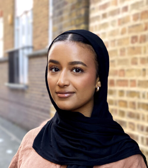 A woman wearing a black hijab and light brown top stands in front of a brick wall, looking at the camera with a slight smile—capturing the approachable spirit often seen in a marketing team. The background is softly blurred.