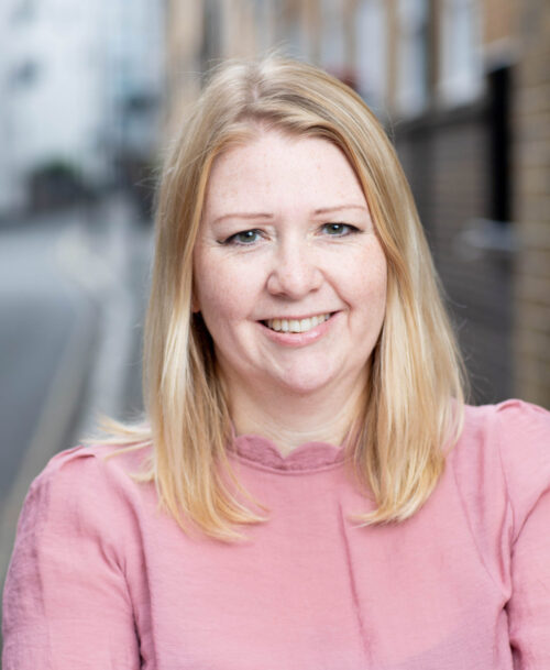 A woman with straight blonde hair and a pink blouse, embodying finance leadership, smiles at the camera while standing on a city street with blurred buildings in the background.