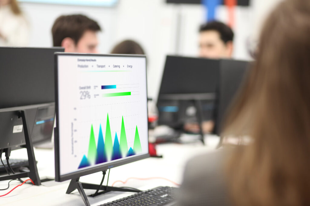 A computer monitor displays a colorful data chart focused on CSR and sustainability, with several people working at desks in the background, slightly blurred. The screen highlights graphs and statistics related to responsible business practices.