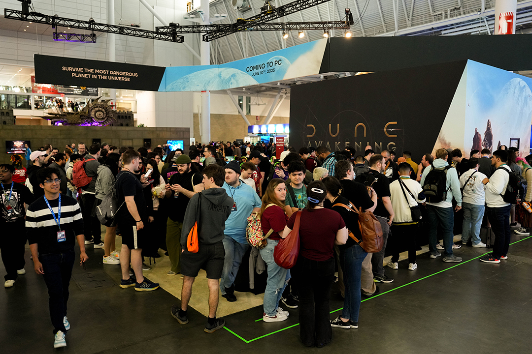 A large crowd gathers in front of a black booth with “Dune Awakening” signage at a gaming convention. People are waiting in line, many wearing backpacks and badges, with colorful banners and lights above.