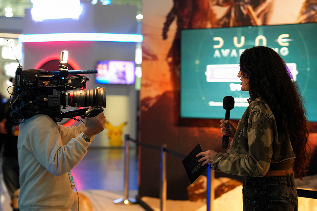 A woman holding a microphone is interviewed on camera at an event, standing in front of a large screen displaying Dune Awakening. The scene appears to be inside a convention center.