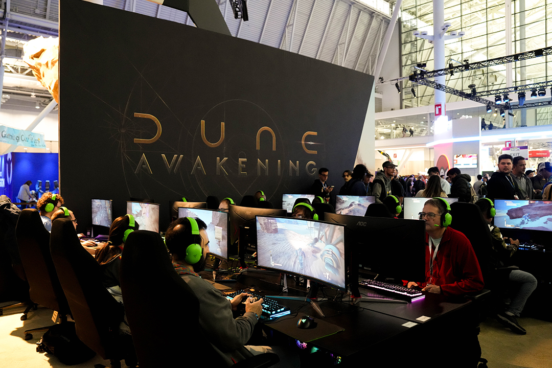 A group of people wearing green headphones play on computers at a gaming event in front of a large wall displaying the Dune: Awakening logo. Spectators stand behind them in a brightly lit convention hall.