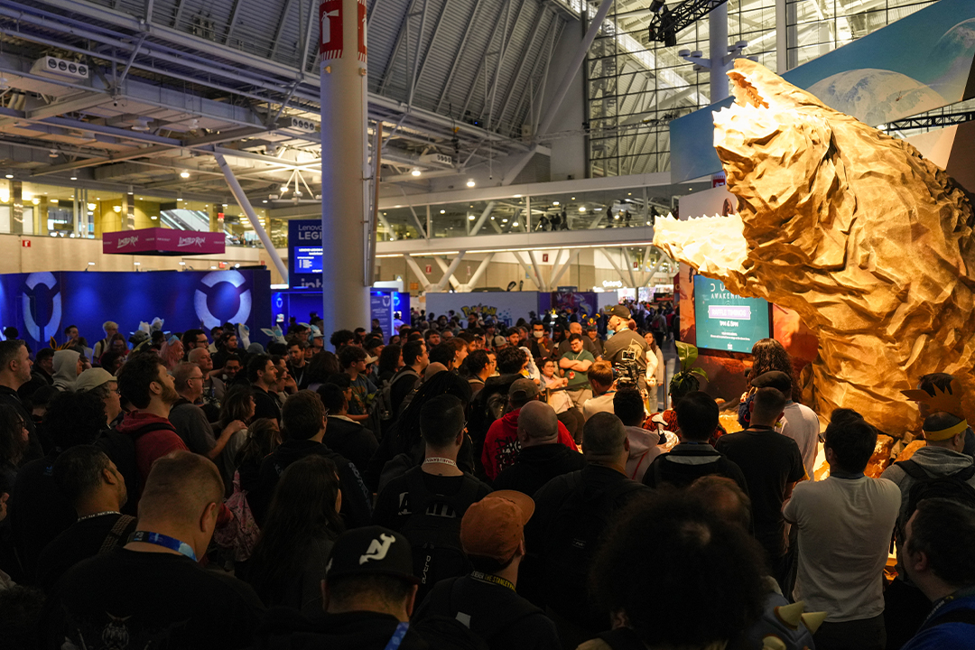 A large crowd gathers at an indoor convention, watching an event near a giant sculpted bear head with its mouth open. Colorful lights and booths are visible in the background.