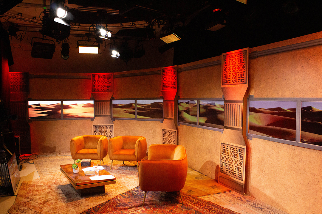 A brightly lit TV studio set features three mustard-yellow armchairs, a low table with books and bottles, and wall panels showing desert dunes under blue sky, with warm red and orange accents throughout.