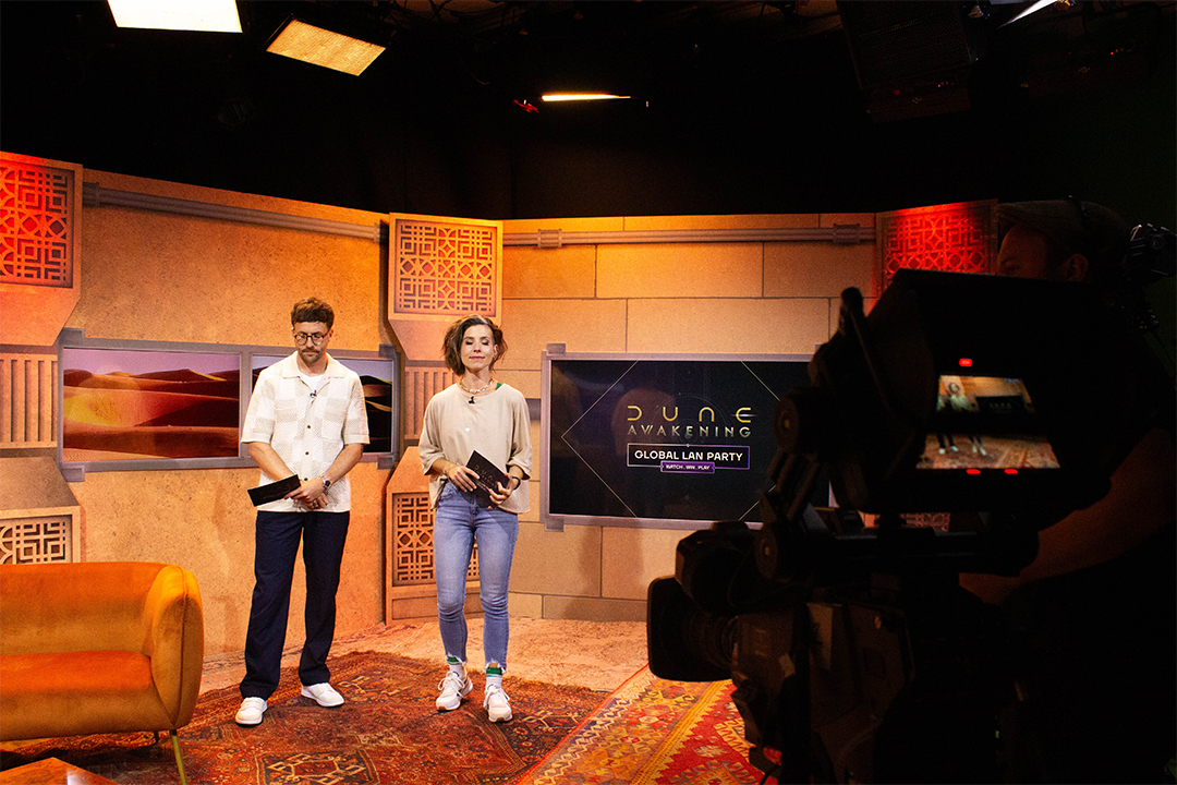 Two hosts stand in a studio with warm lighting, holding notes. A Dune: Awakening Global LAN Party graphic is displayed on a screen behind them. A camera operator films the scene, and desert-themed decor is visible.