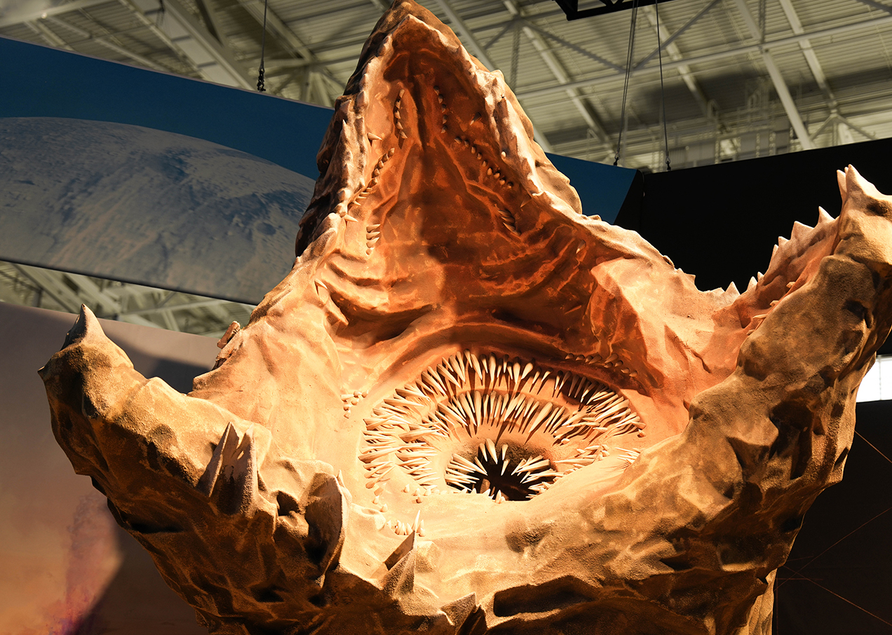 A large, detailed sculpture of a sandworm’s open mouth, showing sharp, concentric teeth and a rough, textured outer surface, displayed indoors under bright lights.