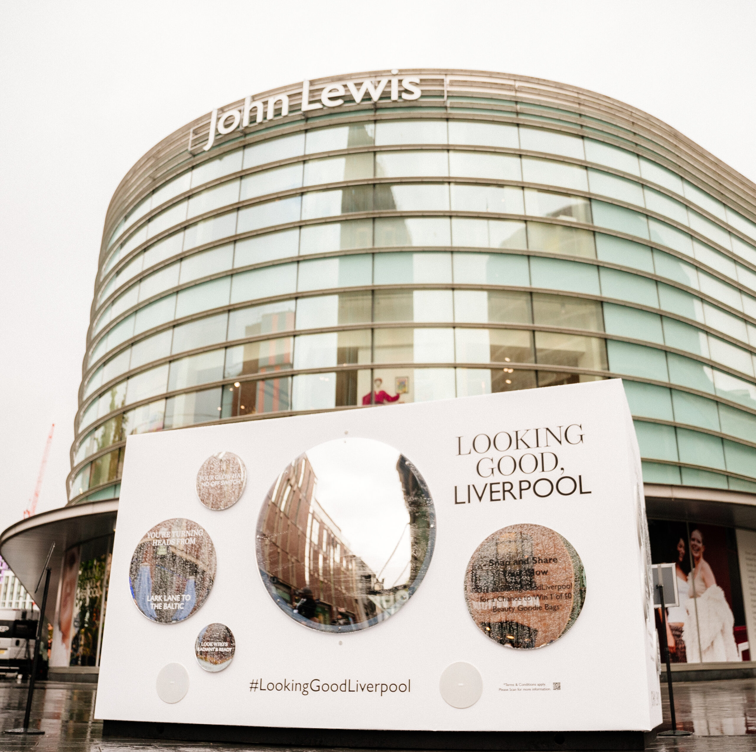 A large curved John Lewis building in Liverpool stands behind a white display board with round mirrors and the text “LOOKING GOOD, LIVERPOOL” and the hashtag #LookingGoodLiverpool on a rainy day.