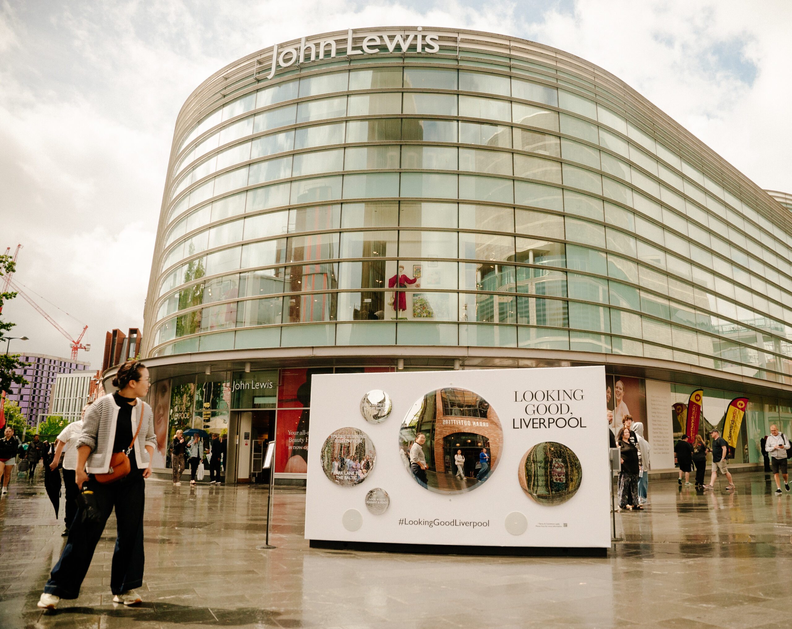 A woman walks past a modern glass-fronted John Lewis store in Liverpool. In front of the building is a display board reading LOOKING GOOD, LIVERPOOL with photos and circular mirrors on a wet pavement.