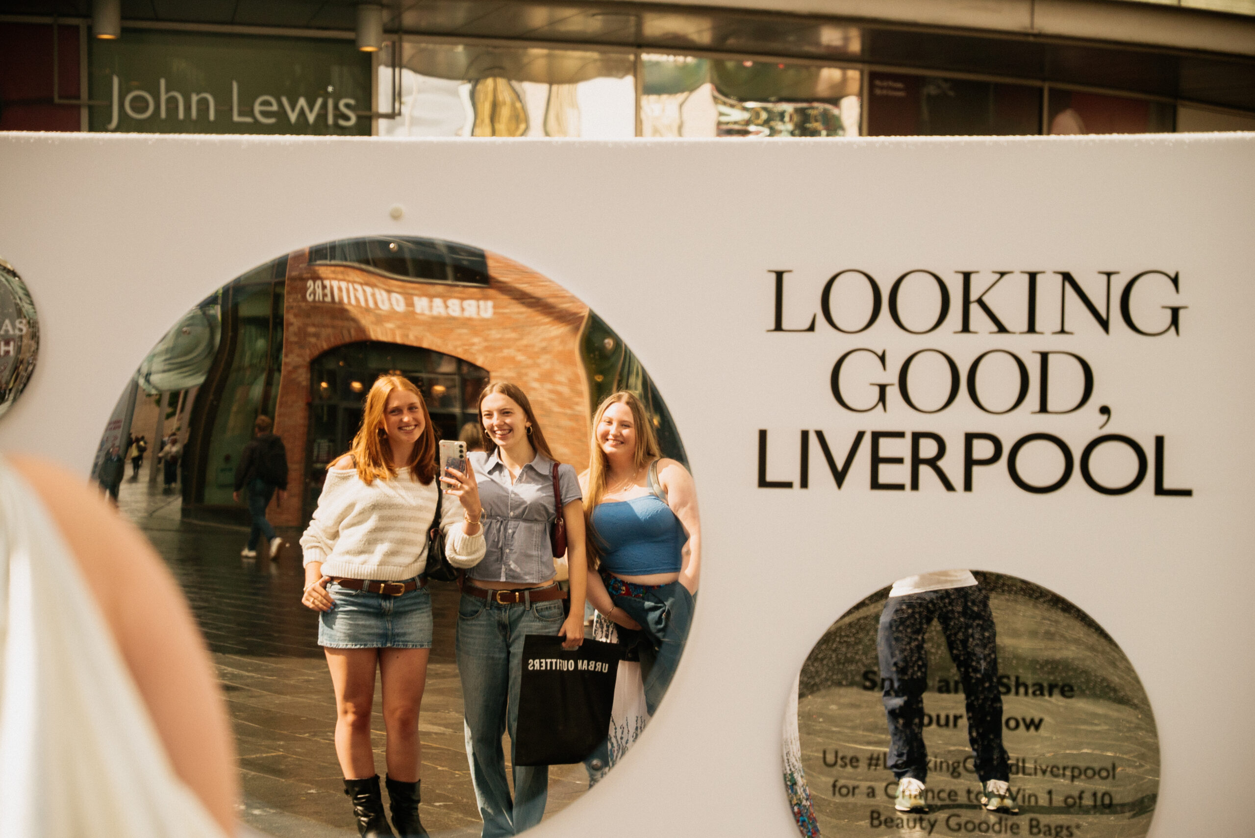 Three smiling young women pose for a selfie in a circular mirror on a city street. The mirror is part of a display that says LOOKING GOOD, LIVERPOOL in bold letters. Urban Outfitters and John Lewis stores are visible.