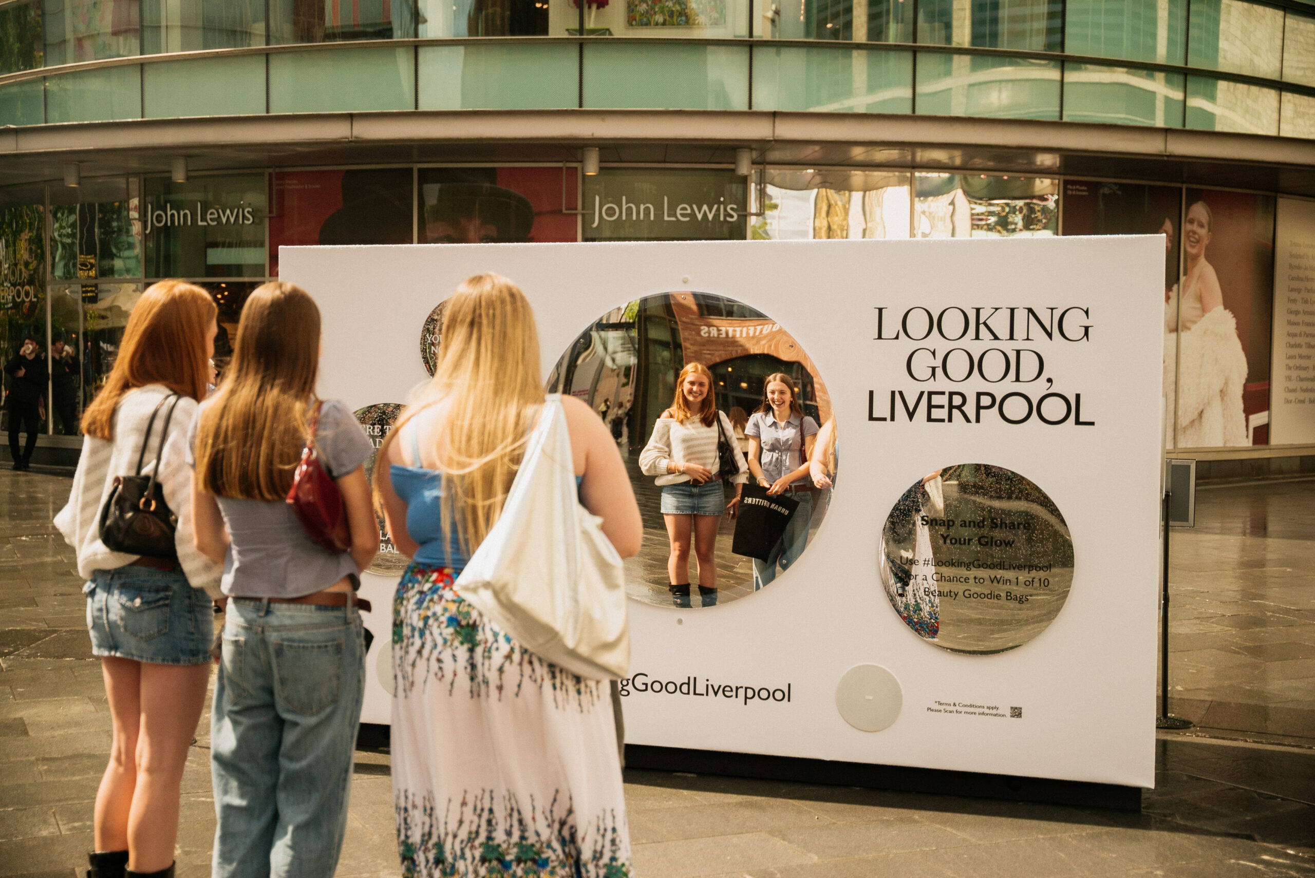 Four young women stand outdoors, looking at their reflection in a large public mirror installation with the text LOOKING GOOD, LIVERPOOL displayed above it, outside a John Lewis store.