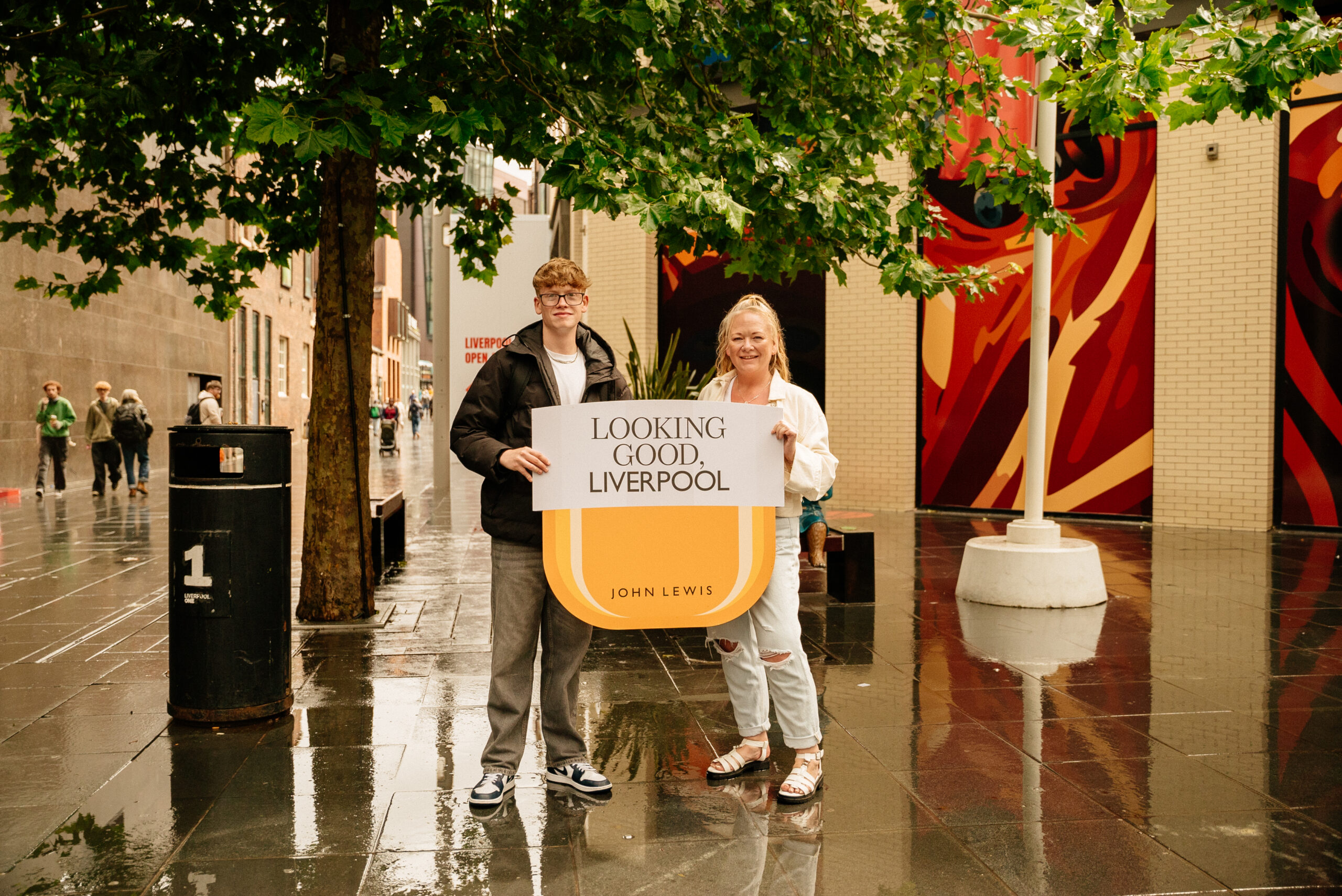 Two people stand on a wet city street, holding a large sign that says “Looking Good, Liverpool” with “John Lewis” beneath. They are smiling, and a colorful mural and green trees are visible in the background.
